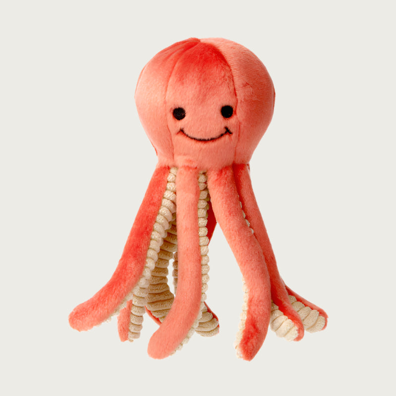 Plush Squirt Octopus dog toy from Fluff & Tuff. Its body is coral colored, with the underside of its legs a light beige with ribbed texture. It has a cute smiley face (eyes & mouth) embroidered in black on its head.