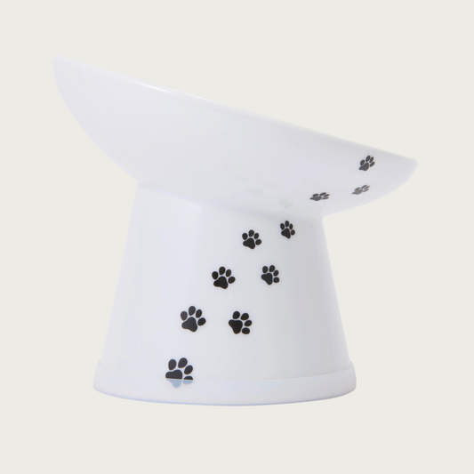 Side view of the tilted, raised, white porcelain cat food bowl with black paw print design trailing up the side.
