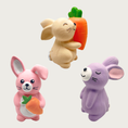 Load image into Gallery viewer, Trio of small, standing bunny-shaped latex dog toys. One on bottom left is pink with a folded ear, holding a carrot. One in top center is light beige with closed eyes, holding a large carrot. One on bottom right is light purple with closed eyes.