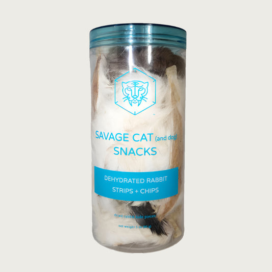 A 3 oz clear plastic container, filled with dehydrated rabbit strips and chips (rabbit hide pieces) by Savage Cat. Fur covered pieces in various natural shades from brown to white are visible.