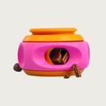 Load image into Gallery viewer, A colorful rubber dog puzzle toy, with a bright pink rotating "dial" on an orange base, with a round opening in front, which can be covered by spinning the pink "dial" part. Treat pieces can be seen inside an scattered around the front of the toy.