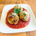 Load image into Gallery viewer, Three golden-brown breaded "arancini" balls served on a bed of red tomato sauce on a white square plate, garnished with grated cheese and basil and parsley leaves, set on a wooden table.