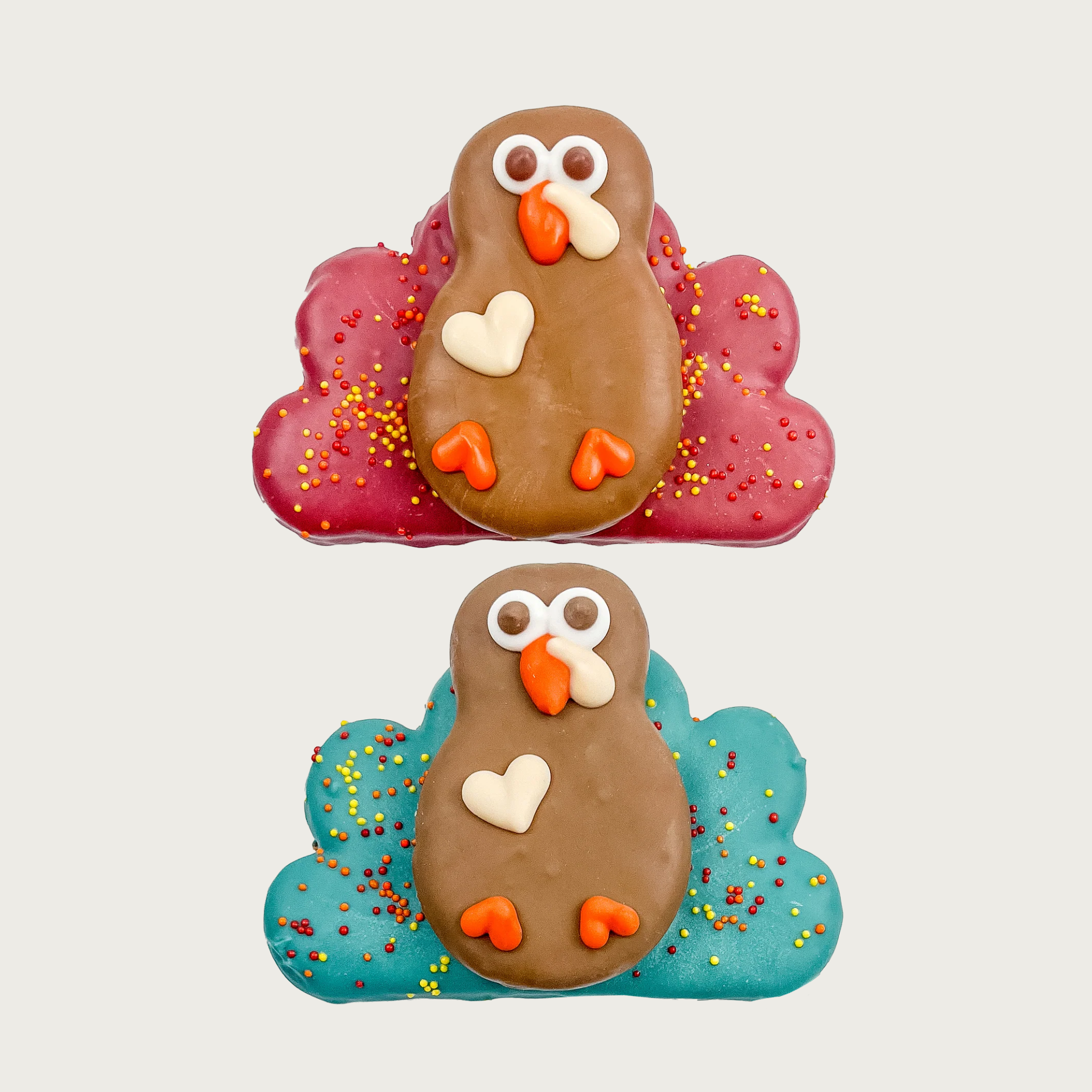 Two decorated dog cookies shaped like turkeys. Both have brown bodies, and piped eyes, beaks & feet. They are stacked cookies - the body is sitting on top of another cookie that's shaped like either red or teal feathers, with multicolored nonpareils.