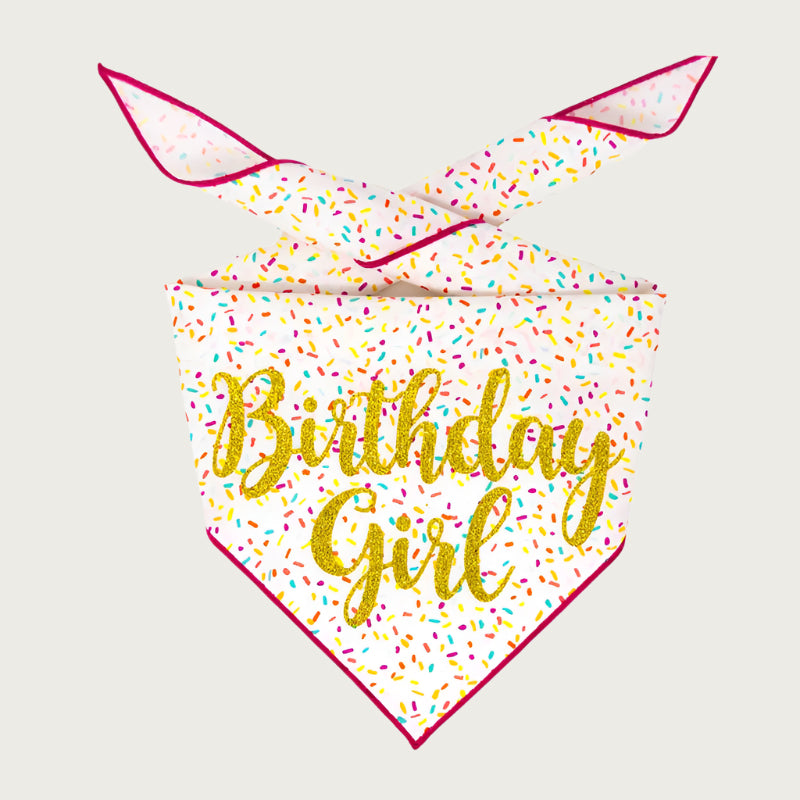 A pet bandana with a white base and multicolored sprinkles pattern and pink trim, featuring the script text 'Birthday Girl' in gold glitter.