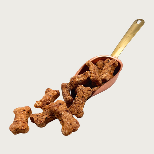 A copper scoop full of Breakfast Omelette Bones treats, that are also spilling out in front of it. The individual treats are medium brown in color with dark flecks, and have a traditional dog bone shape.