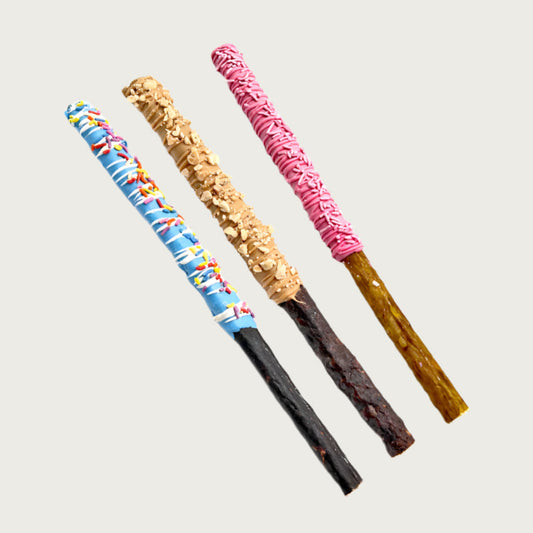 A sample of three decorated Dip Stick dog treats. One is dipped halfway in a blue coating, drizzled with white, with multi-color sprinkles. The middle treat is dipped halfway in peanut butter coating and sprinkled with chopped peanuts. The last treat is dipped halfway in a medium pink coating, drizzled with pink, with pale pink sprinkles.