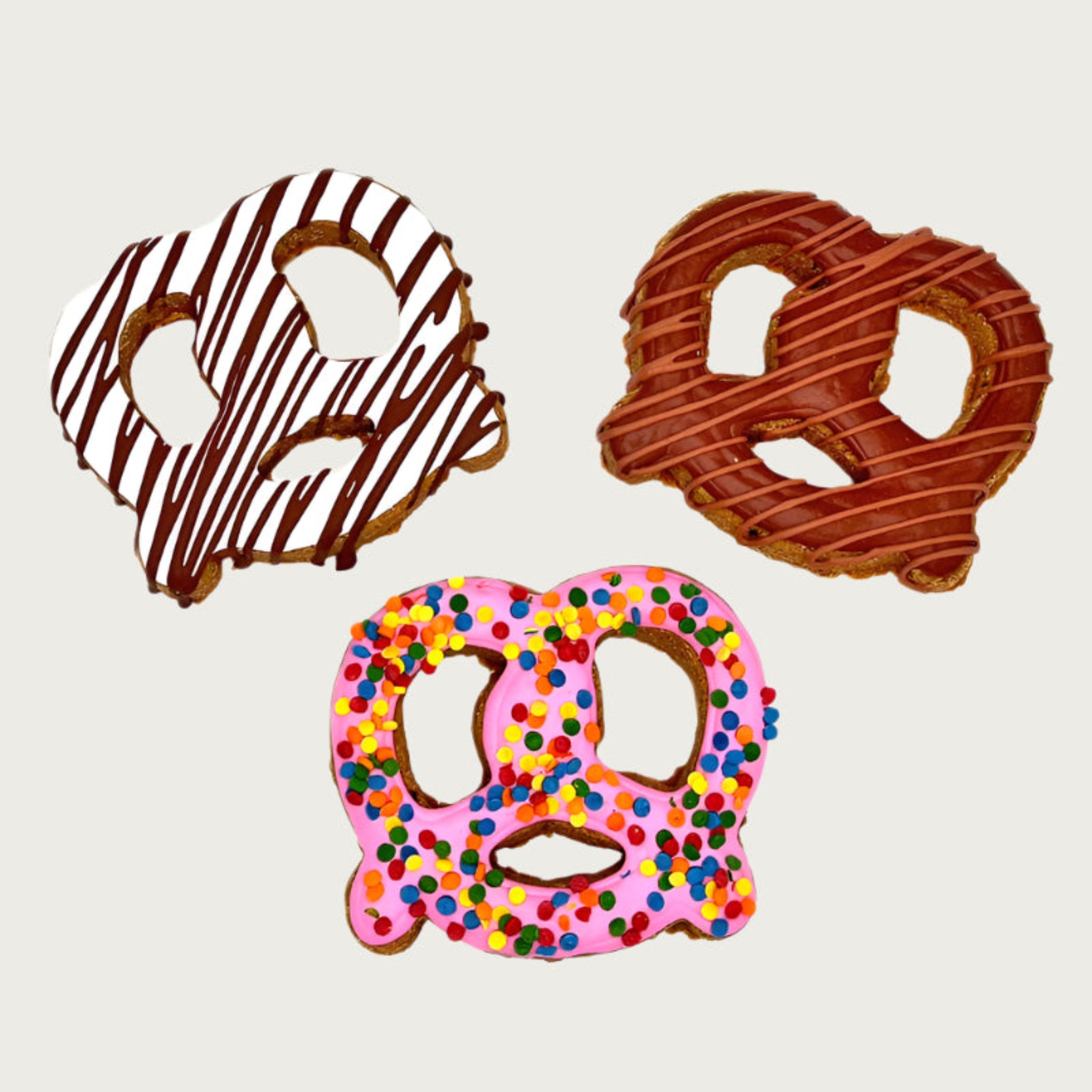 Three pretzel-shaped dog cookies with different toppings; white with brown drizzle, brown with tan drizzle, pink with confetti sprinkles.