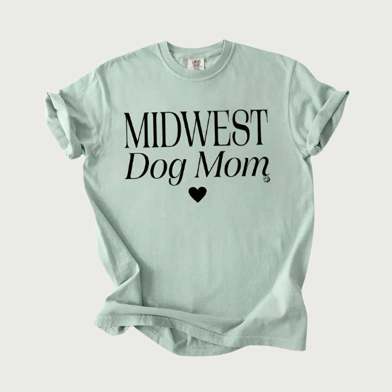 Sage green short sleeved t-shirt with crew neck, with "Midwest Dog Mom" in black text with a small black heart underneath. "Midwest" is in all caps, and "Dog Mom" is slightly italicized.