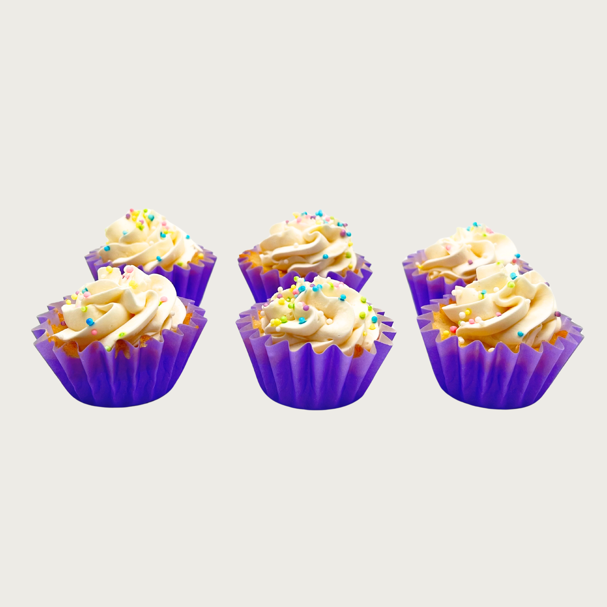 Six mini cupcakes with purple wrappers and white frosting with pastel colored sprinkles.