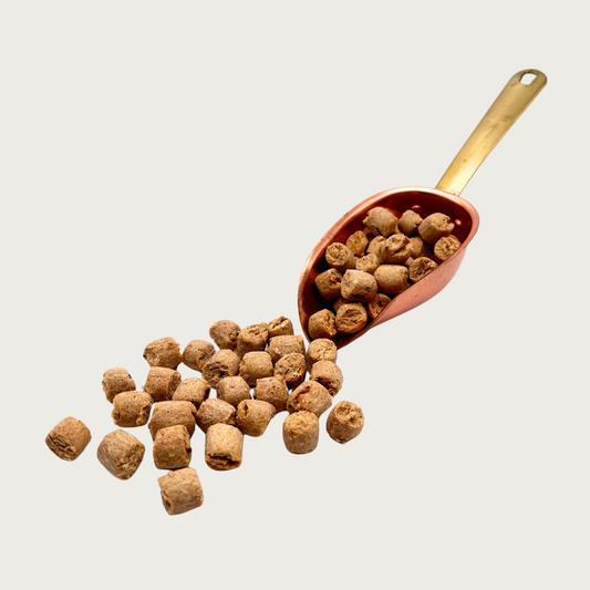 A copper scoop full of Pizza Nibbles, that are also spilling out in front of it. The individual nibbles are tan and about the size and shape of a pencil eraser.