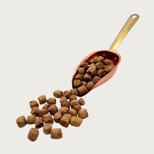 A copper scoop full of Pot Pie Nibbles, that are also spilling out in front of it. The individual nibbles are brown and about the size and shape of a pencil eraser.