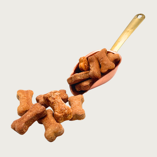 A copper scoop full of Spaghetti & Meatball treats, that are also spilling out in front of it. The individual treats are medium brown in color and have a traditional dog bone shape.
