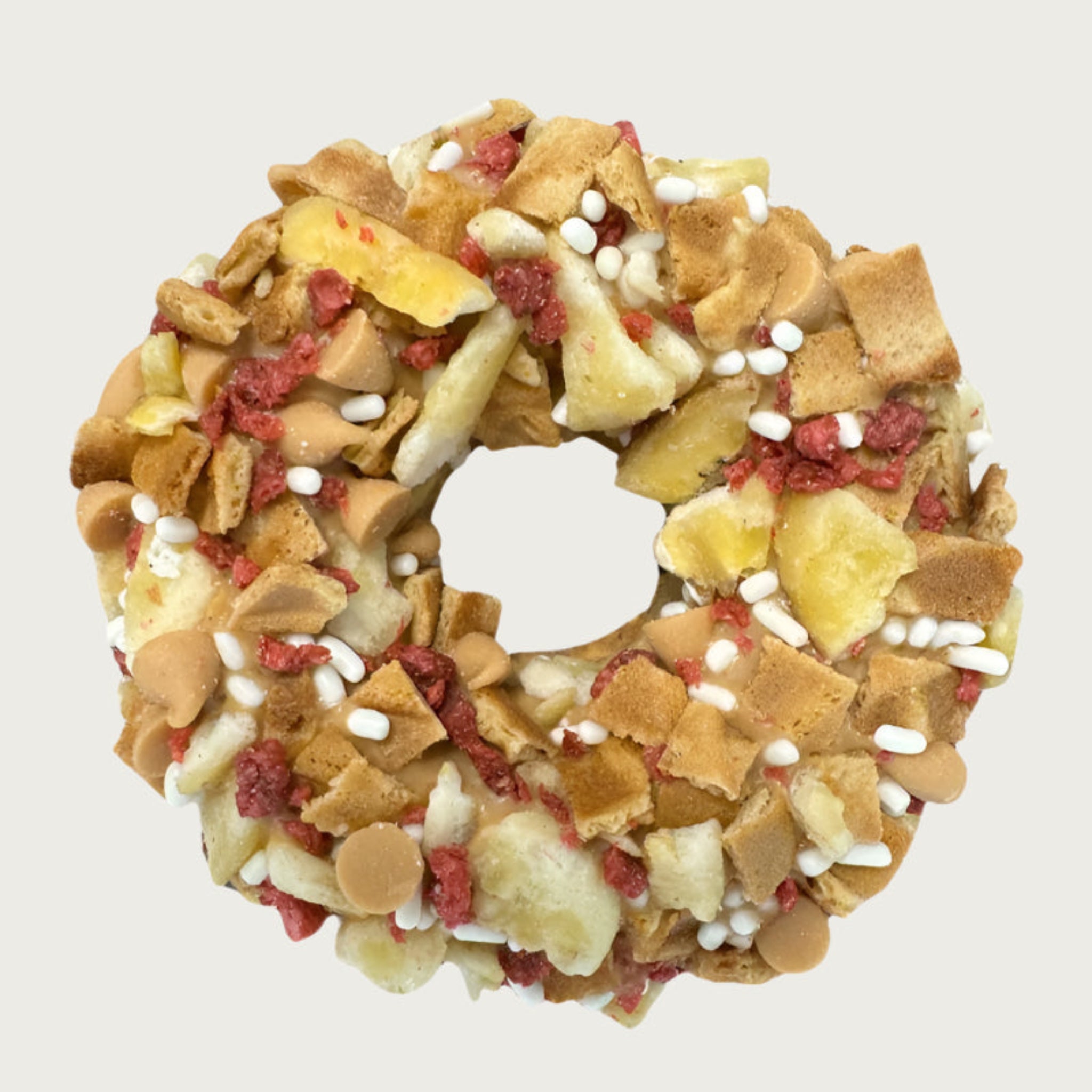Thick, donut-shaped dog cookie decorated with bacon bits, dried banana bits, mini peanut butter drops, waffle cone pieces, and white sprinkles.