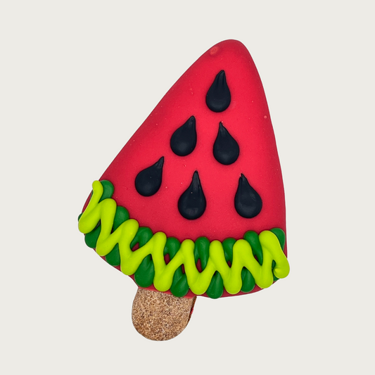 Decorative dog cookie shaped like a watermelon slice with red fruit, black seeds and green rind, with an undecorated part that looks like a popsicle stick.
