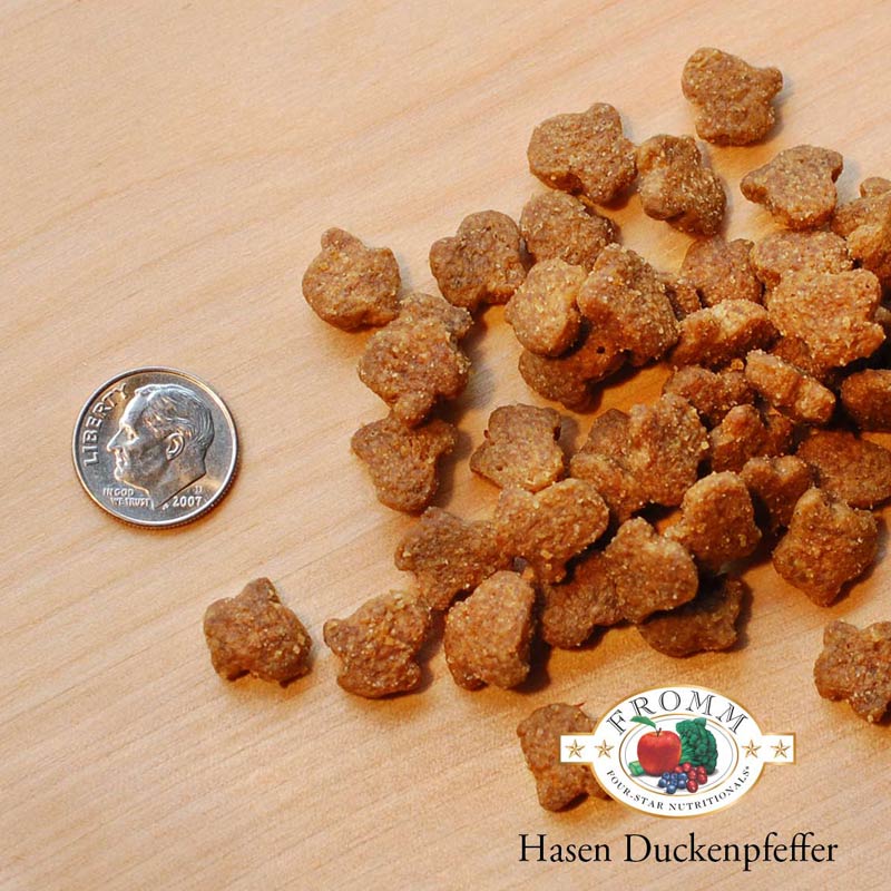 A close-up image of a handful of dry Hasen Duckenpfeffer kibble pieces, next to a dime for size comparison. 2 of the pieces would fit into the area of the dime. They have a slightly rounded animal shape to them.