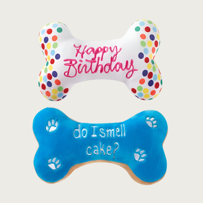 Two styles of birthday bone dog toys, side by side. Top bone is white with multicolored "confetti" circles on the ends, with "Happy Birthday" written in pink script in the center. Bottom bone is bright blue with white pawprints in each corner (for a total of 4), and lower-case white text in center reads "do I smell cake?".
