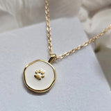 Simple gold chain necklace with a subtly irregular, round shaped pendant. Pendant is trimmed in gold, with a small gold paw print at the center. Pendant background is creamy white enamel.