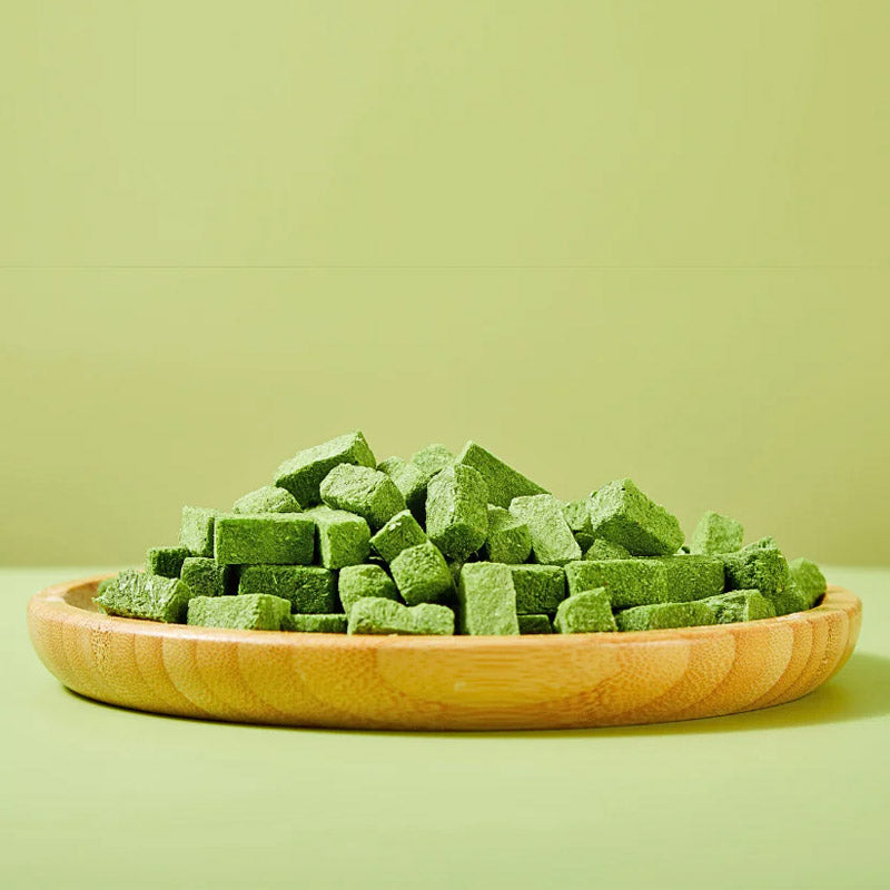 Closeup, side view of Arya! Sit Freeze-Dried Cat Grass with Chicken bites piled onto a wooden plate. They are green, square to rectangular shaped cubes, about 1/2" to 1" long.