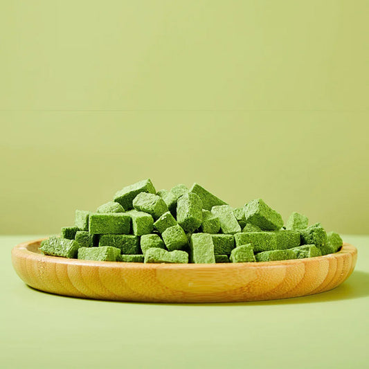 Closeup, side view of Arya! Sit Freeze-Dried Cat Grass with Chicken bites piled onto a wooden plate. They are green, square to rectangular shaped cubes, about 1/2" to 1" long.