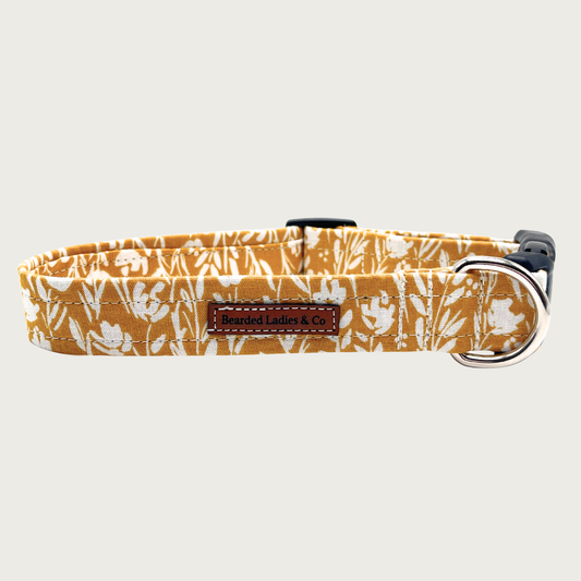Dog collar with mustard yellow background and abstract/boho style cream floral print, with black plastic hardware and silver metal D-ring.