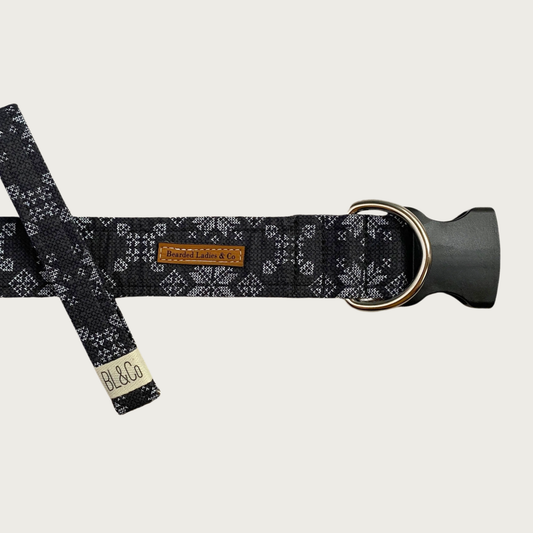 Dark gray dog collar featuring a light cross-stitched style snowflake pattern in white. It has a black plastic clip and silver toned D-ring. A smaller, thinner collar in the same design is leaning on the left side of the larger one, which is the focal point of the image.