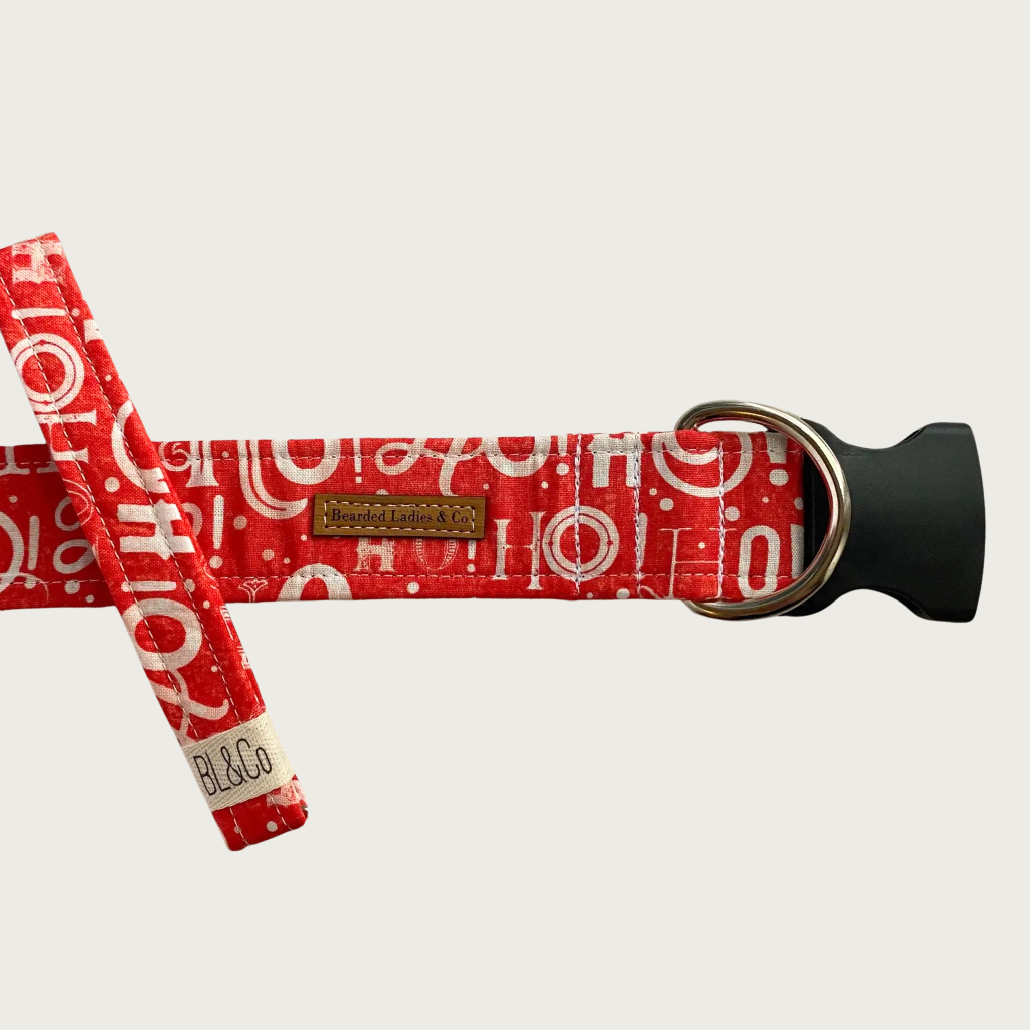 Christmas-themed red dog collar featuring the expression "Ho-ho-ho!" in various white fonts. It has a black plastic clip and silver toned D-ring. A smaller, thinner collar in the same design is leaning on the left side of the larger one, which is the focal point of the image.