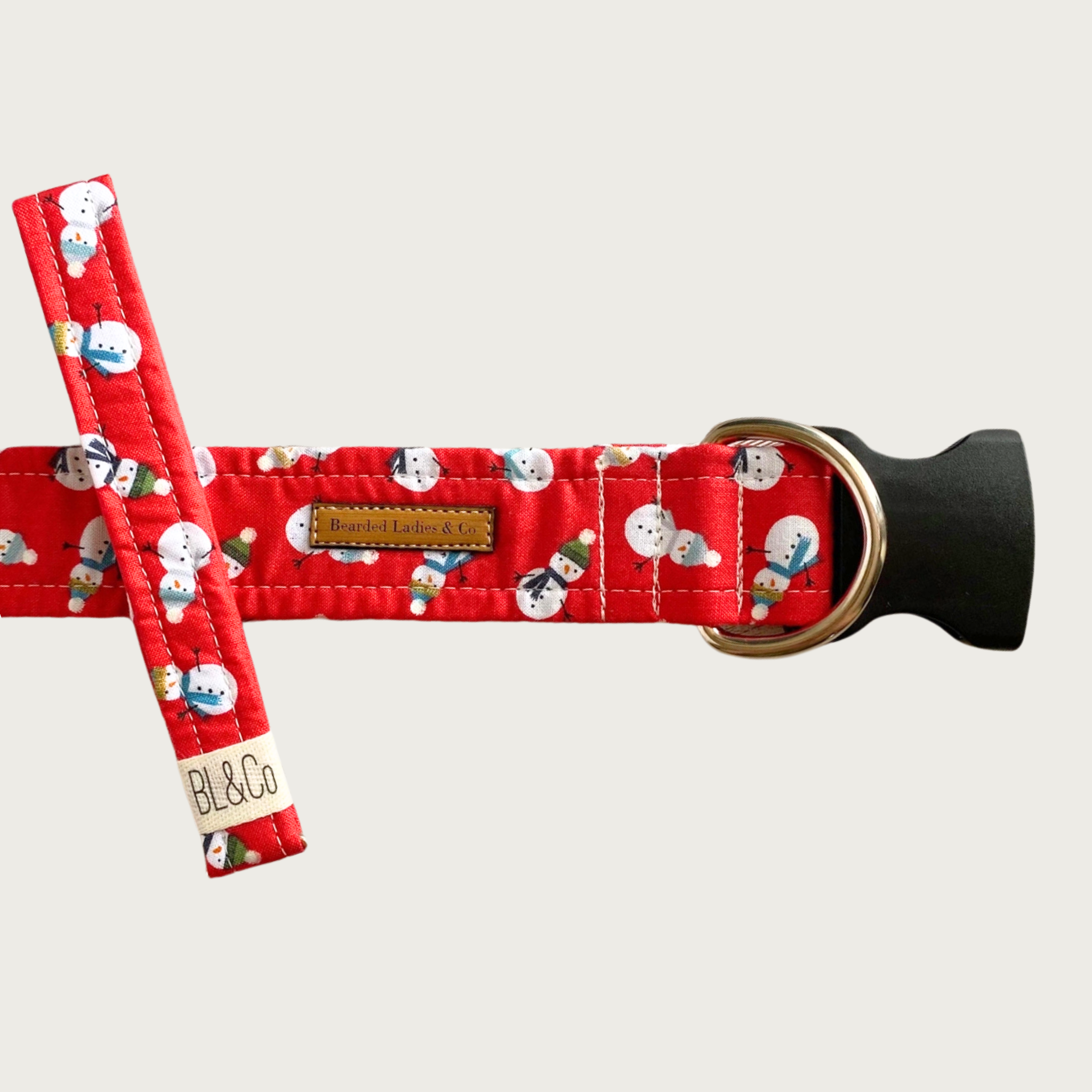 Winter-themed red dog collar with a mini snowman print. The snowmen are wearing different colored hats & scarves. It has a black plastic clip and silver toned D-ring. A smaller, thinner collar in the same design is leaning on the left side of the larger one, which is the focal point of the image.