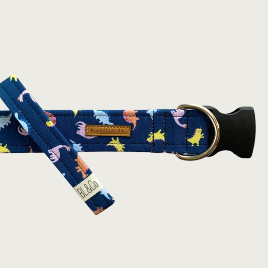 Navy blue dog collar with mini pastel-colored dinosaurs of different kinds. It has a black plastic clip and silver toned D-ring. A smaller, thinner collar in the same design is leaning on the left side of the larger one, which is the focal point of the image.