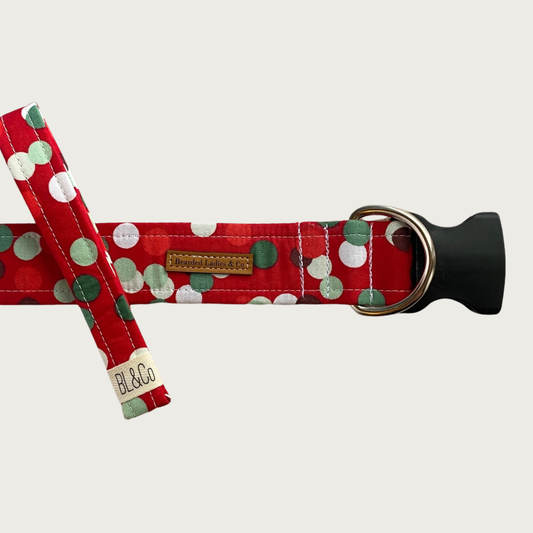 Christmas-themed red dog collar with green, white, and red polka dots in a randomly scattered pattern. It has a black plastic clip and silver toned D-ring. A smaller, thinner collar in the same design is leaning on the left side of the larger one, which is the focal point of the image.