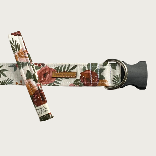 Floral patterned dog collar featuring an antique-style print of roses in mauve tones and greenery in a deep shade of green. It has a black plastic clip and silver toned D-ring. A smaller, thinner collar in the same design is leaning on the left side of the larger one, which is the focal point of the image.