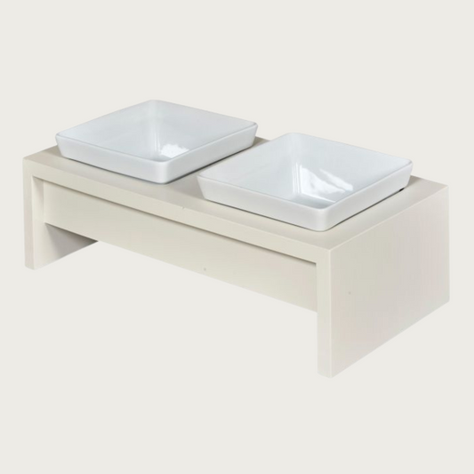 Double bowl, elevated dog feeder. Stand is a creamy white color, in a simple rectangular design. Square white ceramic bowls in center.