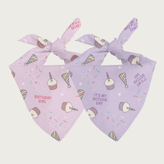 Two sides of the same dog bandana, shown side by side. Both feature the same imagery-illustrated cupcakes and party hats with tiny white paw prints and multi colored streamers on the background. On the left is the pink side that has "Birthday Girl" text, on the right is the light purple side that has "It's My Gotcha Day" text.