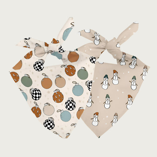Two sides of the same dog bandana, shown side by side. One side has a cream background with simple Christmas ornaments in muted shades of rusty orange and blue-green. The other side has a beige background with simple white snowmen wearing similar hued stocking caps.