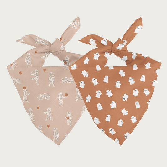 Two sides of the same dog bandana, shown side by side. One side has a pinkish-cream background with simple mummies in different actions. The other side has a light burnt orange background with simple white ghosts in different actions.