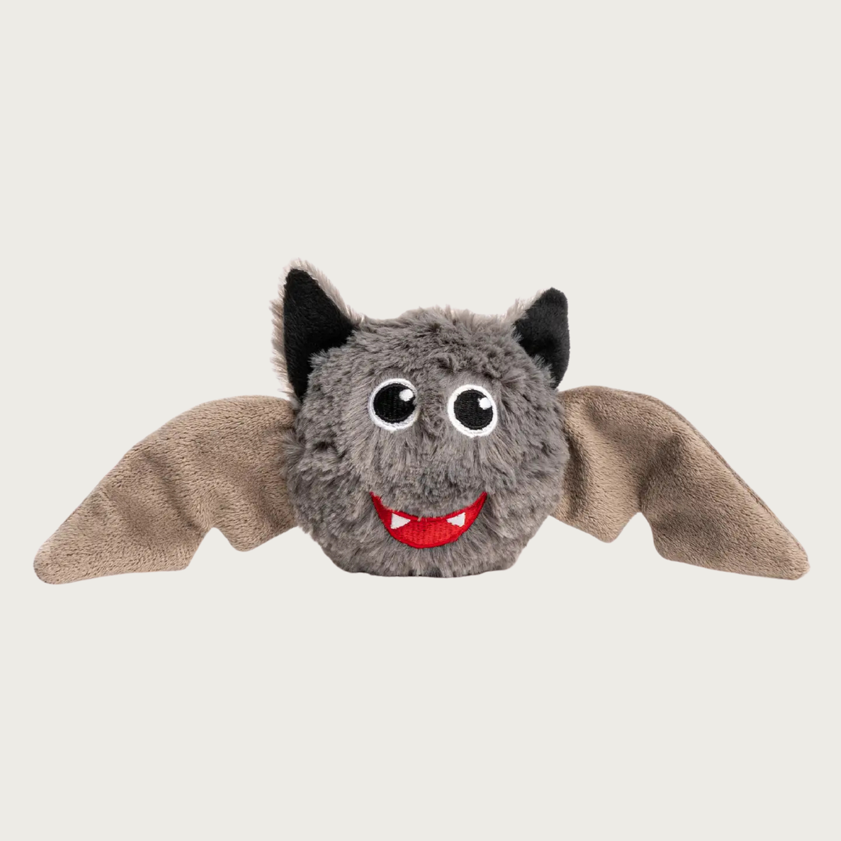 Plush dog toy shaped like a bat with outstretched wings. Its body is a shaggy gray plush, with tan microplush wings, and black plush ears. It has embroidered black & white eyes and a smile with white fangs on a red background.