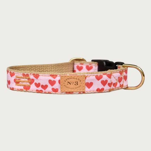 Valentine's themed dog collar. Base color is pastel pink, with a pattern of simple reddish pink hearts. Underside of collar is light tan. Has brass hardware and black plastic clip.