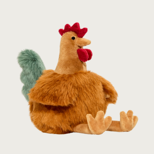 Plush chicken dog toy. It has a tan, round, plump body, with short wings and dangling legs, and grayish-green colored tail. Its head/neck features red comb and wattle, and soft beak. 