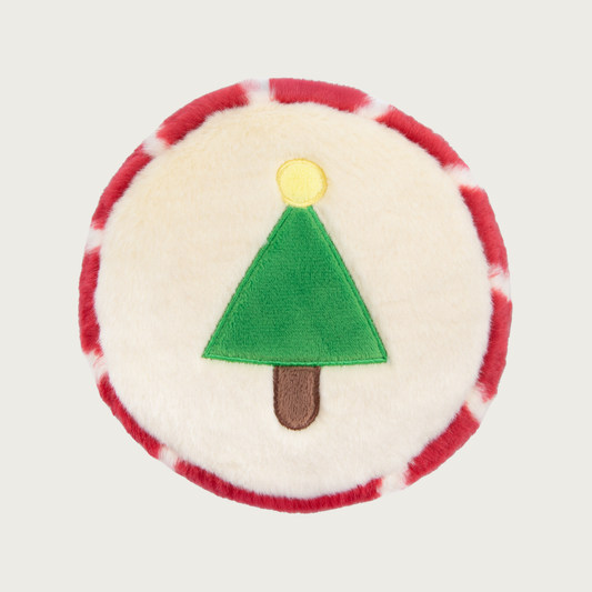 Christmas-themed, round plush dog toy. In the center is a simple green Christmas tree with brown trunk and yellow circle "star" on top. The body of the toy is cream colored, and the edge of the toy is red & cream striped.