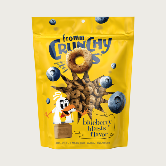 A yellow pouch of Fromm Crunchy Os blueberry blasts flavor dog treats. Mini photos of  whole blueberries are featured, along with an illustration of a cartoon chicken pushing down on the handle of an old-time dynamite blaster, directed at the center window which shows the brown, ring shaped treats.