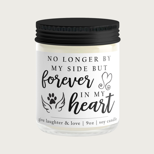 Clear glass candle jar with a black lid and white label, with text that reads "No longer by my side, but forever in my heart" in a combination of script and serif fonts. Illustrated imagery includes angel wings with a paw print in center, and script heart. Candle wax is a natural ivory color. 9 oz.