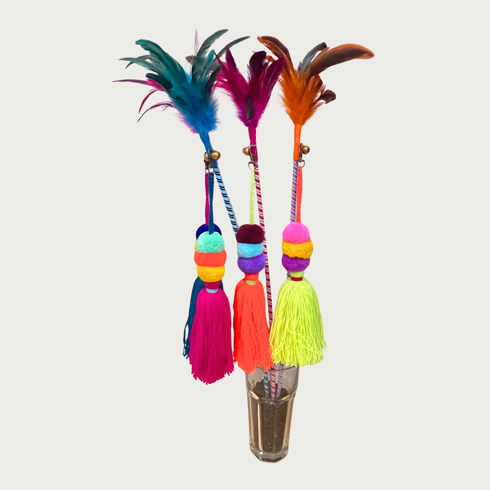 Four multicolored cat teaser wands propped up in a glass. They have feathers and a small bell on one end, and a large brightly colored pom pom tassel attached beneath the feathers. 