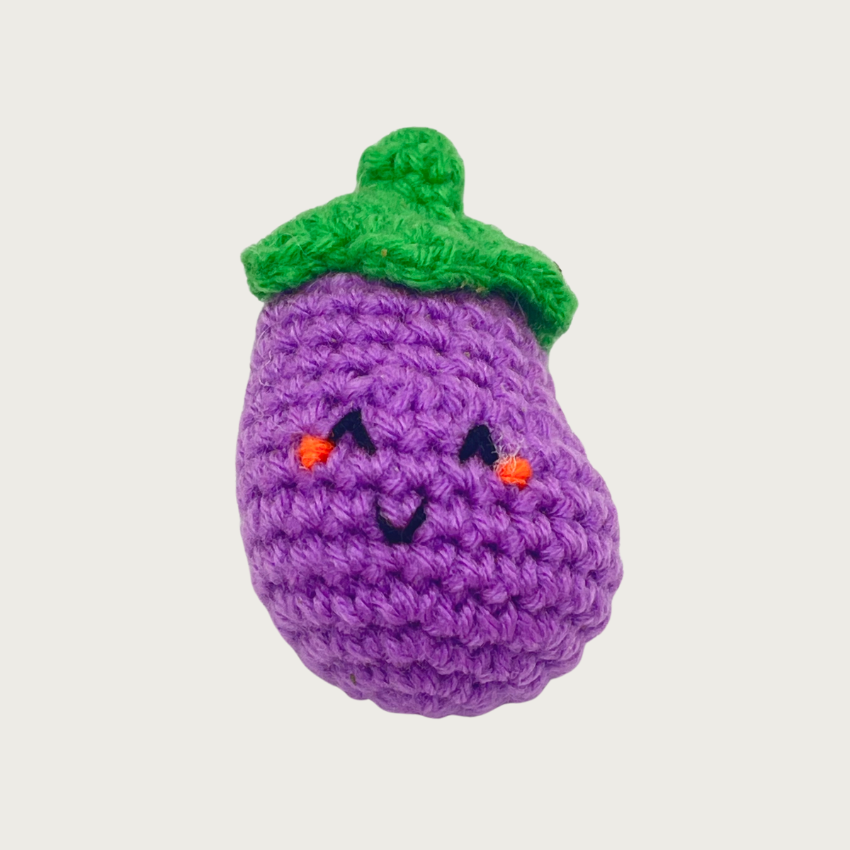 A crocheted eggplant cat toy with a purple body, green stem, and embroidered smiling face.