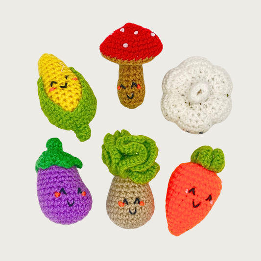 Six crocheted vegetable shaped cat toys arranged in two rows: on top is an ear of corn, mushroom, and head of garlic. Beneath that is an eggplant, turnip, and carrot.