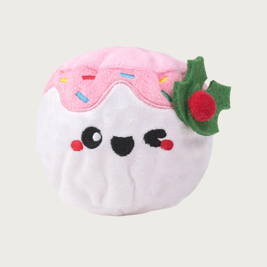 Christmas-themed ball shaped dog toy. Its plush exterior cover is white, with an embroidered smiley facial expression. It's topped with a pale pink plush "frosting" with multicolored embroidered sprinkles, and felt holly sprig accent.