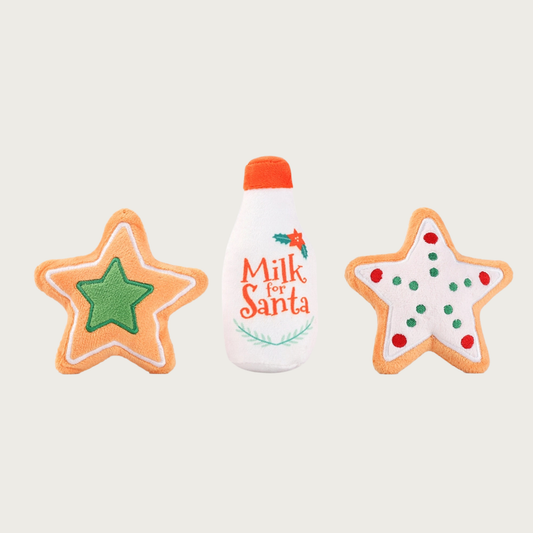 Group of three Christmas-themed plush dog toys. There are two star-shaped cookies, one colored white with red & green dot accents, and the other with a small green star in the center, and a line outline along the star's edges. In the center is a bottle-shaped toy labeled 'Milk for Santa' with decorative holly & evergreen accents. It is white with a red "cap".