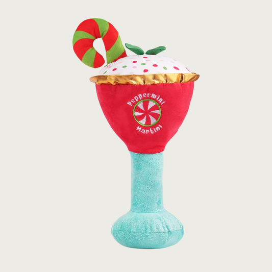 Plush, Christmas-themed dog toy resembling a peppermint martini cocktail. It has a light blue stem, red "bowl" trimmed with gold fabric, white plush "drink" inside with multicolored dots, and a red & green candy cane "garnish". On the front is an embroidered round peppermint candy, with text that reads "Peppermint Martini".