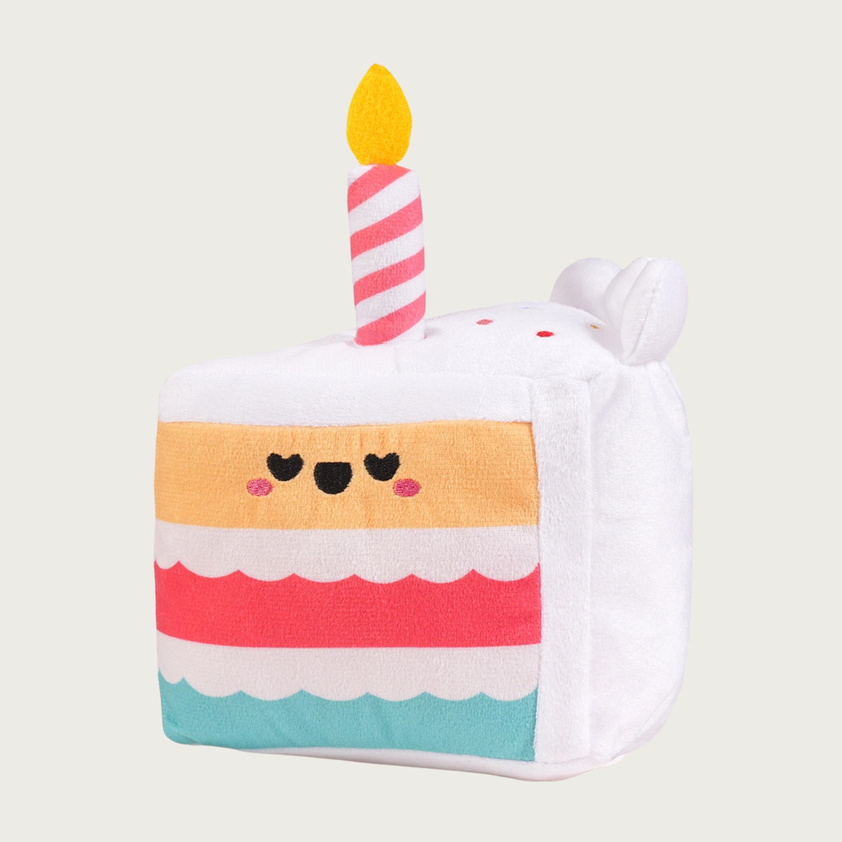 A plush dog toy shaped like a slice of birthday cake. It has 3 multi colored layers with white "frosting" in-between them, and white frosting on the top and back of the cake. The top cake layer has an embroidered smiley face. On top is a pink & white striped, plush birthday candle with a yellow "flame".