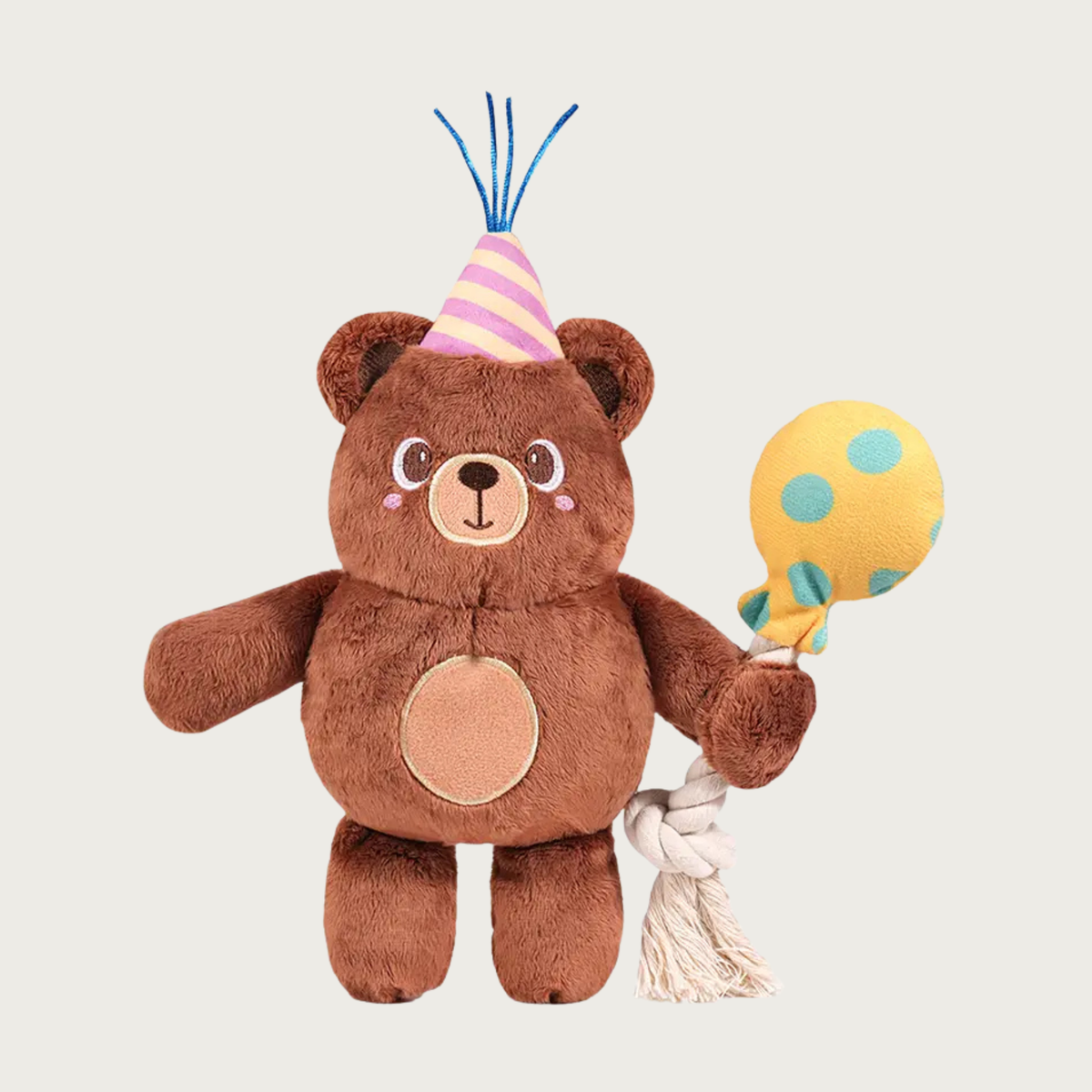 A birthday party themed, plush teddy bear dog toy. The bear is medium brown with light brown tummy & snout, and cute embroidered face. It's wearing a pink & peach diagonally striped party hat with blue sparkly ribbon sticking out. The bear is holding a neutral colored, cotton rope balloon; the plush balloon is yellow with teal polka dots.