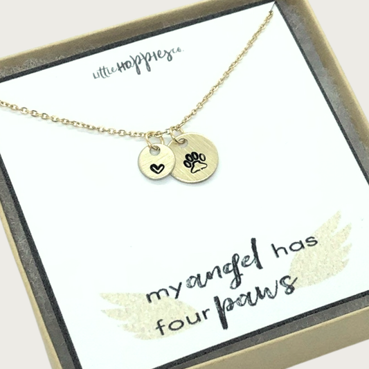 Gold-toned, delicate chain link necklace with small, round charm with outlined black paw print detail, and a smaller coordinating charm with an outlined heart. Set on white card that reads "my angel has four paws" with angel wings accent. Comes in brown gift box.