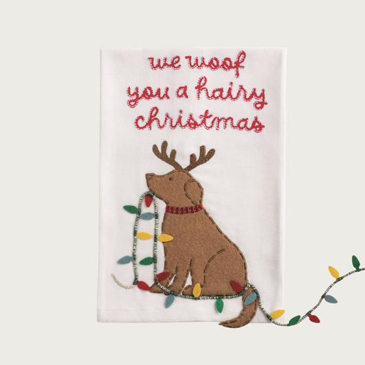 Christmas-themed white decorative towel, with a tan labrador-type dog wearing reindeer antlers and holding a string of holiday lights in its mouth, that trail off the bottom of the towel. Red script text on top reads: "We woof you a hairy Christmas".